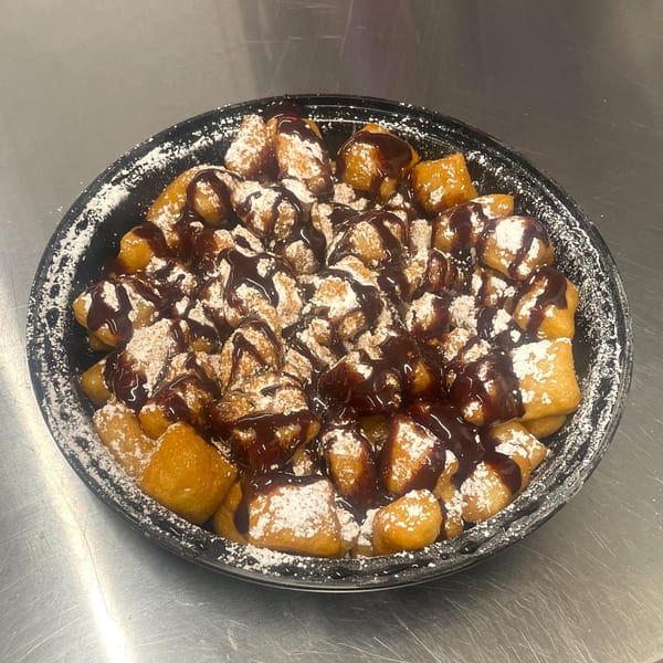 A black bowl filled with golden-brown fried dough pieces, drizzled with chocolate sauce and powdered sugar.