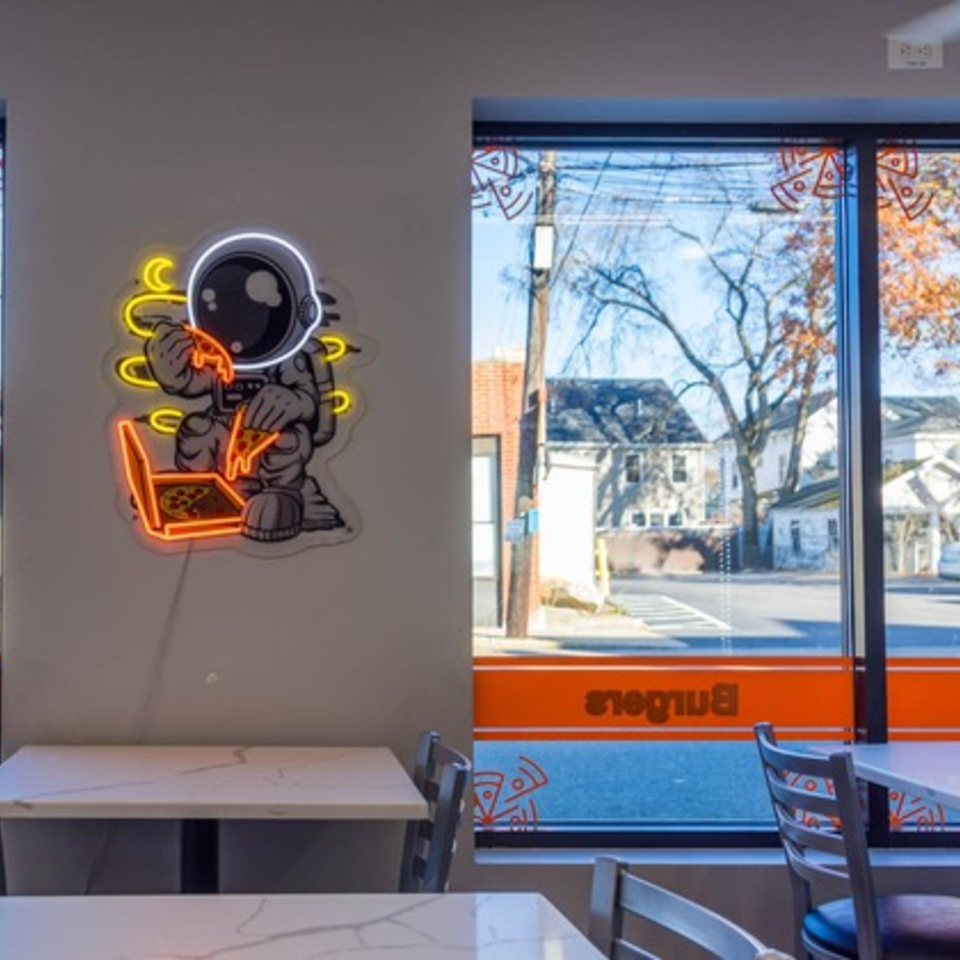 Neon astronaut eating pizza at Johnny's Pockets, with tables and a window view of the street.