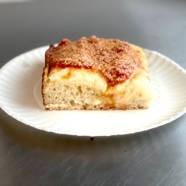 A square slice of Johnny's Pockets pizza with a thick crust and melted cheese on a white paper plate.